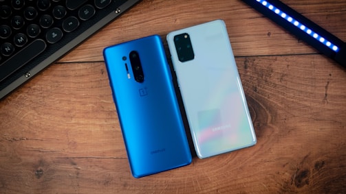 Two smartphones are placed on a wooden surface. The phone on the left is blue with a vertical camera setup, and the one on the right is a reflective, light-colored phone with a rectangular camera module. Nearby, part of a keyboard and an LED light strip are visible on the sides.