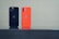 Two smartphones are standing upright on a reflective black surface. One phone is black with dual rear cameras, while the other is orange with dual cameras as well. Both have a company logo on the back.