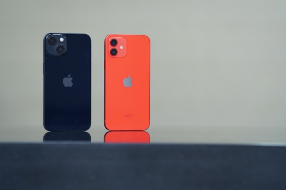 Two smartphones are standing upright on a reflective black surface. One phone is black with dual rear cameras, while the other is orange with dual cameras as well. Both have a company logo on the back.