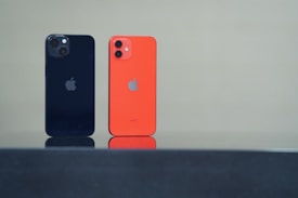 Two smartphones are standing upright on a reflective black surface. One phone is black with dual rear cameras, while the other is orange with dual cameras as well. Both have a company logo on the back.