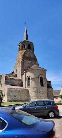 A historic stone church with a tall spire and arched windows stands prominently against a clear blue sky. In front of the church, two cars are parked, one blue and one gray. The church features various architectural details, including a circular apse and a steeply pitched roof.