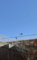 A brick building is shown with a metal railing along the top edge. The sky is clear and blue, and two security lights are mounted on poles near the roofline.