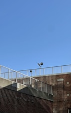 A brick building is shown with a metal railing along the top edge. The sky is clear and blue, and two security lights are mounted on poles near the roofline.