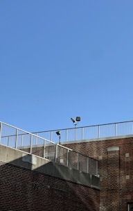 A brick building is shown with a metal railing along the top edge. The sky is clear and blue, and two security lights are mounted on poles near the roofline.