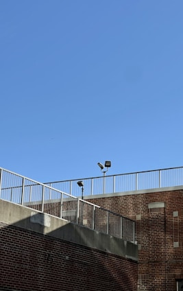 A brick building is shown with a metal railing along the top edge. The sky is clear and blue, and two security lights are mounted on poles near the roofline.