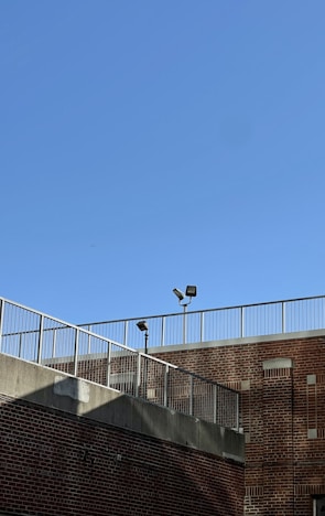 A brick building is shown with a metal railing along the top edge. The sky is clear and blue, and two security lights are mounted on poles near the roofline.