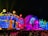 A vibrant festival stage features a large, colorful sculpture of an owl with glowing eyes and outstretched wings. The structure is illuminated with a kaleidoscope of lights in blue, red, and green hues, creating an energetic atmosphere. A crowd of people is gathered in front, suggesting a lively event or concert.
