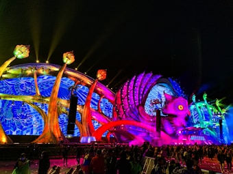 A vibrant festival stage features a large, colorful sculpture of an owl with glowing eyes and outstretched wings. The structure is illuminated with a kaleidoscope of lights in blue, red, and green hues, creating an energetic atmosphere. A crowd of people is gathered in front, suggesting a lively event or concert.