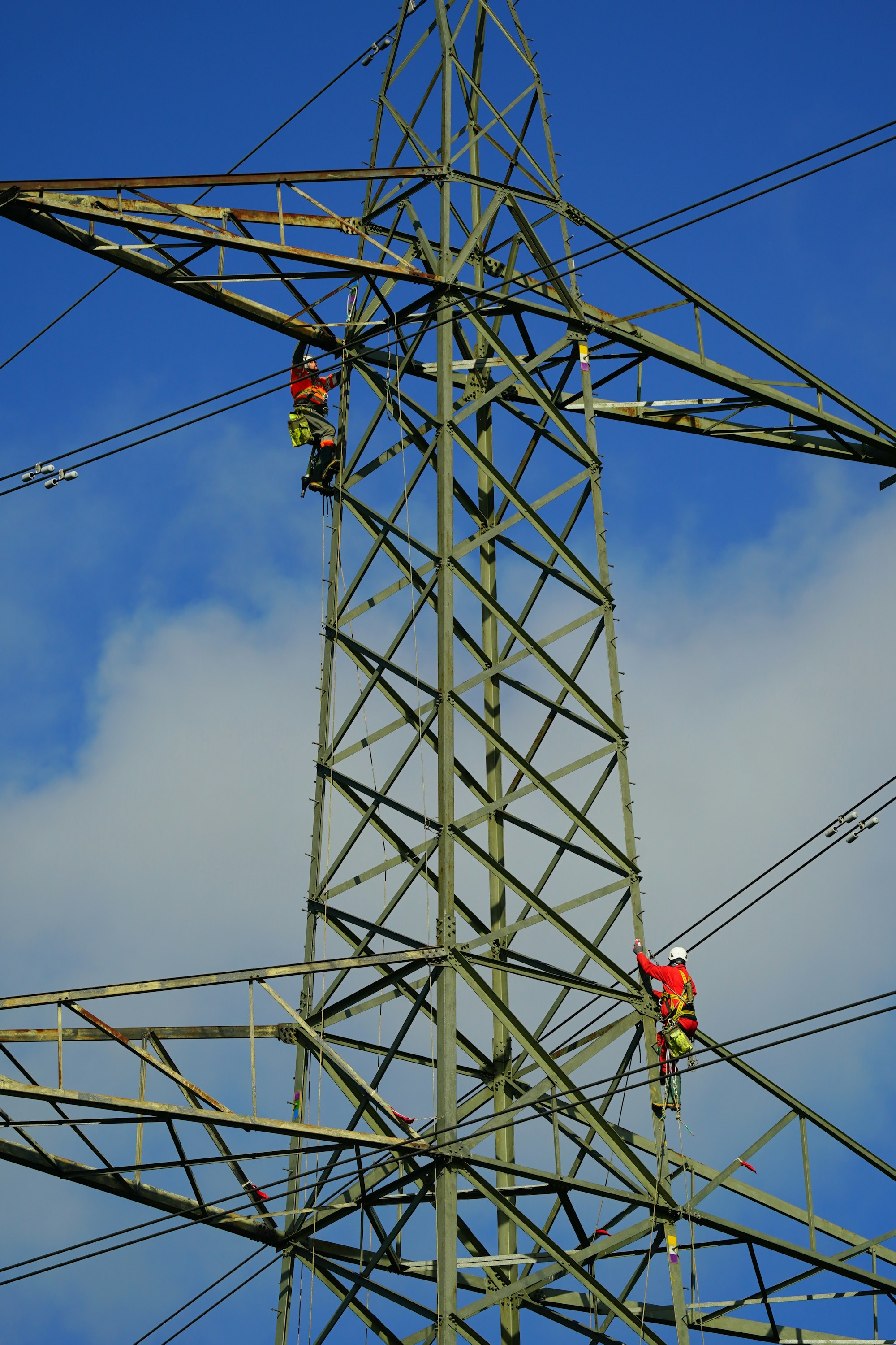 Two workers in high visibility clothing and climbing harnesses partway up a steel lattice transmission tower, with conductors running off to either side against a blue sky.