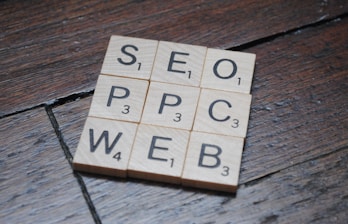 Wooden Scrabble tiles on a dark wooden surface form a grid with letters spelling SEO, PPC, WEB. Each tile has a letter with a small number indicating its point value in the game.