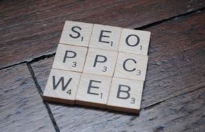 Wooden Scrabble tiles on a dark wooden surface form a grid with letters spelling SEO, PPC, WEB. Each tile has a letter with a small number indicating its point value in the game.