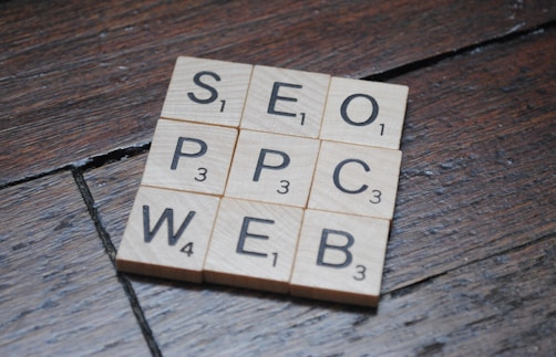 Wooden Scrabble tiles on a dark wooden surface form a grid with letters spelling SEO, PPC, WEB. Each tile has a letter with a small number indicating its point value in the game.