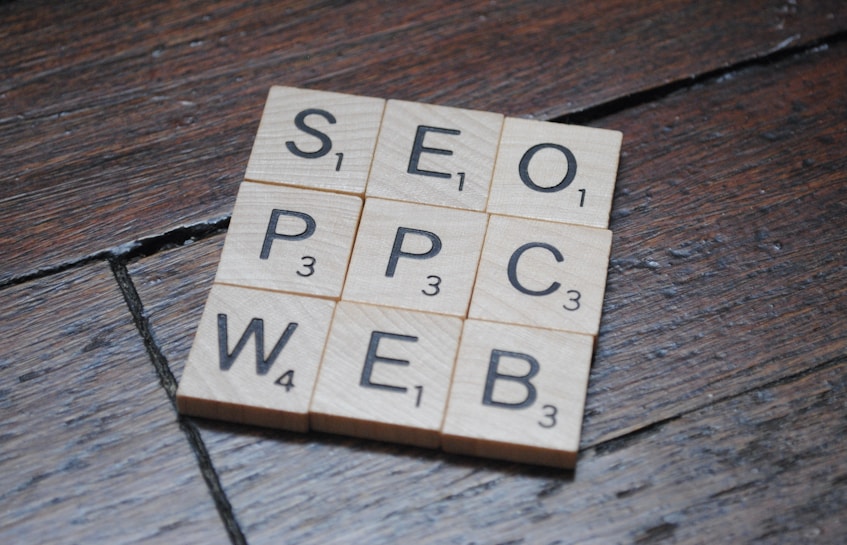 Wooden Scrabble tiles on a dark wooden surface form a grid with letters spelling SEO, PPC, WEB. Each tile has a letter with a small number indicating its point value in the game.