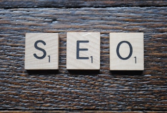 The image contains three Scrabble tiles placed side by side on a wooden surface. The tiles display the letters 'S', 'E', and 'O', each with a value of one point.