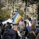 A large group of people gathered outdoors, with a prominent rainbow flag displaying the word 'PAIX'. The setting includes tall trees and a historic building in the background. People appear engaged, possibly in a demonstration or peaceful rally. Various other smaller flags and signs are visible.