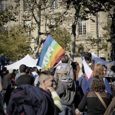 A large group of people gathered outdoors, with a prominent rainbow flag displaying the word 'PAIX'. The setting includes tall trees and a historic building in the background. People appear engaged, possibly in a demonstration or peaceful rally. Various other smaller flags and signs are visible.