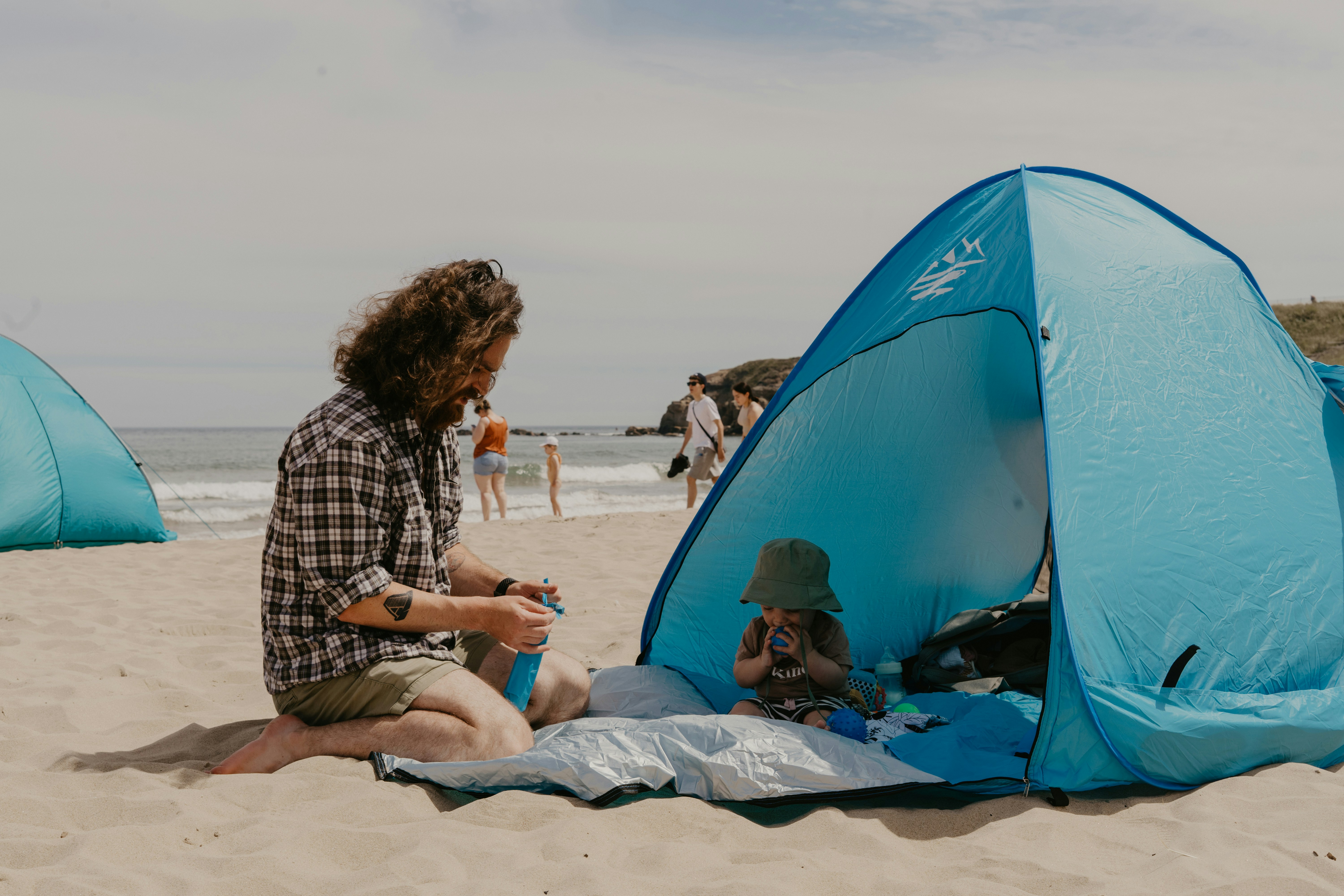 Man in a plaid shirt sitting on the sand beside a blue pop-up beach tent where a small child in a green sun hat sits in the shade, sea and other beachgoers behind