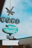 A vintage-style sign with the word 'BOWL' at the top, followed by signs for 'Cocktail Lounge', 'Billiards', 'Arcade', and 'Coffee Shop'. The signs are predominantly blue with a slightly worn appearance and are set against a clear sky.