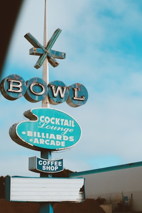 A vintage-style sign with the word 'BOWL' at the top, followed by signs for 'Cocktail Lounge', 'Billiards', 'Arcade', and 'Coffee Shop'. The signs are predominantly blue with a slightly worn appearance and are set against a clear sky.