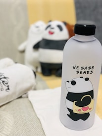 A water bottle in the foreground features a cartoon panda holding a handbag, accompanied by the text 'We Bare Bears.' Behind it are blurry stuffed panda toys arranged on a soft surface.