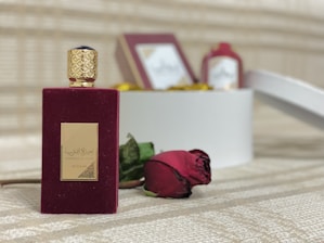 A luxurious dark red perfume bottle with a golden cap is placed on a textured surface. Next to it lies a red rose. In the background, there is a blurred white box with other bottles, creating a soft, elegant setting.