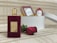 A luxurious dark red perfume bottle with a golden cap is placed on a textured surface. Next to it lies a red rose. In the background, there is a blurred white box with other bottles, creating a soft, elegant setting.
