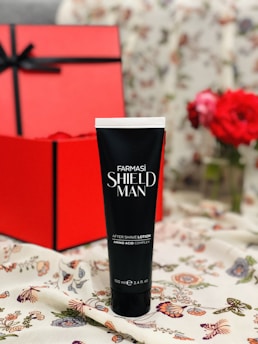 A black tube labeled Farmasi Shield Man After Shave Lotion stands on a floral-patterned fabric. Behind it, a large red gift box tied with a black ribbon is visible. In the background, red flowers add a decorative touch.
