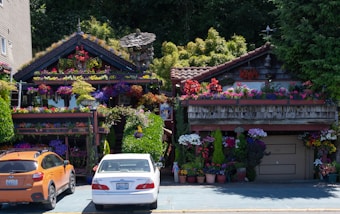 Two houses are adorned with various colorful flowers and plants that cover parts of the structures, creating a lush and vibrant appearance. Each house has a unique design, with one having a thatched roof and the other featuring tile roofing. There are numerous flowerpots at the entrance and bright blooms hanging from the walls and balconies. Two cars, one orange and one white, are parked in front of the houses.