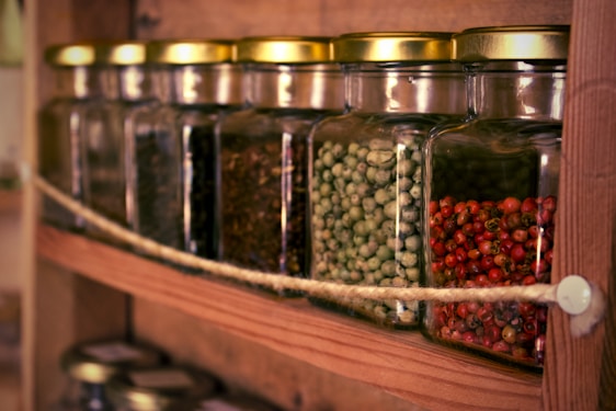 Several glass jars with gold-colored lids are placed in a row on a wooden shelf, containing different types of dried spices or seeds. The jars are secured with a rope across the front.
