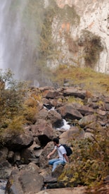 A person wearing a backpack is seen traversing over large rocks in a rugged terrain surrounded by greenery. There is a waterfall cascading down a rocky cliff on the left side, creating mist that blends with the lush foliage. The scene conveys an adventurous outdoor setting, with the individual appearing to hike along a stream flowing between the boulders.
