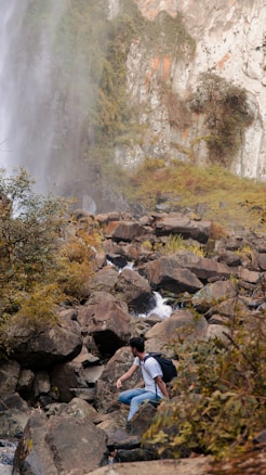 A person wearing a backpack is seen traversing over large rocks in a rugged terrain surrounded by greenery. There is a waterfall cascading down a rocky cliff on the left side, creating mist that blends with the lush foliage. The scene conveys an adventurous outdoor setting, with the individual appearing to hike along a stream flowing between the boulders.