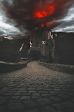A dark and foreboding castle is set against a dramatic red and black sky. The cobblestone path leading to the entrance of the castle gives a sense of ancient mystery and potential danger. The surrounding landscape is shrouded in shadow, enhancing the eerie atmosphere.
