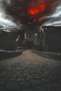 A dark and foreboding castle is set against a dramatic red and black sky. The cobblestone path leading to the entrance of the castle gives a sense of ancient mystery and potential danger. The surrounding landscape is shrouded in shadow, enhancing the eerie atmosphere.