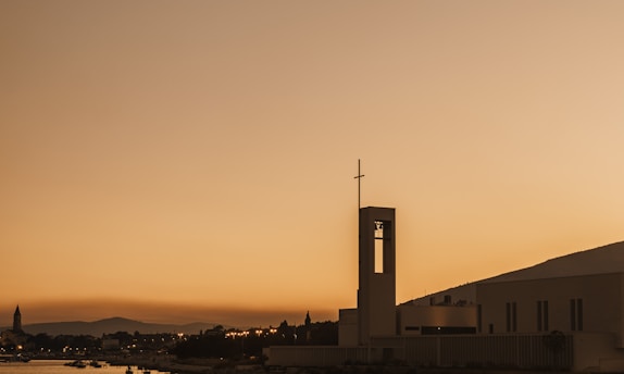 A serene countryside church at sunset with a cross silhouetted against the sky.