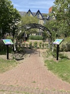 A charming garden pathway leads through an arched metal trellis covered in growing plants, bordered by two signs featuring colorful illustrations. The path is made of red bricks and extends towards a backdrop of lush greenery, trees, and a building with pointed roofs. The scene is set on a sunny day, casting natural light over the garden.