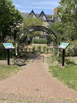 A charming garden pathway leads through an arched metal trellis covered in growing plants, bordered by two signs featuring colorful illustrations. The path is made of red bricks and extends towards a backdrop of lush greenery, trees, and a building with pointed roofs. The scene is set on a sunny day, casting natural light over the garden.