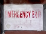 A faded and partially obscured red text spells out 'EMERGENCY EXIT' on a white surface. The sign appears worn, with paint marks and smudges around it on a background that is a mix of brown and gray tones. Small patches of peeling paint are visible.