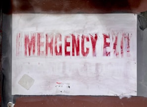 A faded and partially obscured red text spells out 'EMERGENCY EXIT' on a white surface. The sign appears worn, with paint marks and smudges around it on a background that is a mix of brown and gray tones. Small patches of peeling paint are visible.