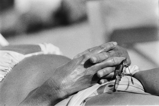 Hands resting on a pregnant belly, held delicately with fingers intertwined. A small pair of glasses can be seen held in one of the hands, suggesting a moment of rest or contemplation. The image is captured in black and white, emphasizing the texture and shadows on the skin.