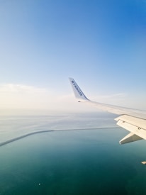 An airplane wing with a Ryanair logo is visible from a window, flying over a vast expanse of calm, blue ocean. The horizon shows a seamless blend of sky and sea, with a gentle curve of a coastline or sea wall visible below.