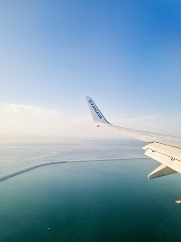 An airplane wing with a Ryanair logo is visible from a window, flying over a vast expanse of calm, blue ocean. The horizon shows a seamless blend of sky and sea, with a gentle curve of a coastline or sea wall visible below.
