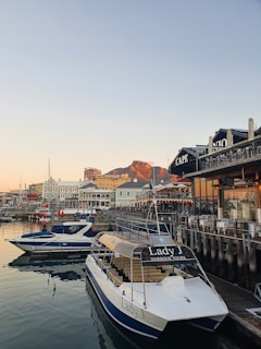 A harbor scene during sunset features several boats docked at the waterfront. A mountain in the background reflects the warm hues of the setting sun. The waters are calm, mirroring the boats and nearby structures. Buildings with nautical themes line the boardwalk, adding to the peaceful maritime atmosphere.