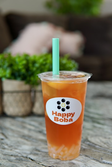 A plastic cup filled with iced tea or boba tea labeled 'Happy Boba' with a green straw. The drink has a lemon slice and appears to have boba or jelly pieces at the bottom. It is placed on a marble or granite surface with a blurred background featuring greenery and soft furnishings.