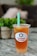 A plastic cup filled with iced tea or boba tea labeled 'Happy Boba' with a green straw. The drink has a lemon slice and appears to have boba or jelly pieces at the bottom. It is placed on a marble or granite surface with a blurred background featuring greenery and soft furnishings.
