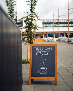 A wooden sandwich board on a sidewalk features a chalkboard with the text 'Tides Café Open' and an illustration of a steaming cup of coffee. The scene is set outdoors with trees and a modern building in the background.