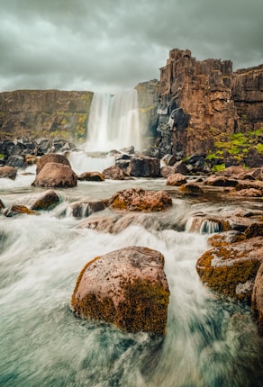 A dramatic waterfall cascades over rugged cliffs into a flowing stream surrounded by large, moss-covered rocks. The sky is overcast, adding to the moody atmosphere of the scene.