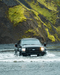 A large black SUV is driving through a shallow body of water, creating splashes around its tires. The background features steep, rugged hills covered with bright green moss and dark rocky surfaces. The scene conveys a sense of adventure and off-road capability.