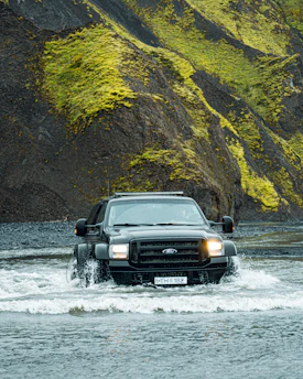 A large black SUV is driving through a shallow body of water, creating splashes around its tires. The background features steep, rugged hills covered with bright green moss and dark rocky surfaces. The scene conveys a sense of adventure and off-road capability.