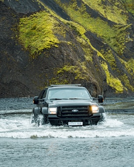 A large black SUV is driving through a shallow body of water, creating splashes around its tires. The background features steep, rugged hills covered with bright green moss and dark rocky surfaces. The scene conveys a sense of adventure and off-road capability.