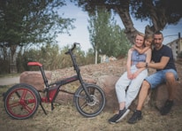 A family of three is sitting on a rock outdoors, surrounded by trees and green foliage. A woman, man, and a child are smiling, displaying a sense of unity and closeness. Beside them is a modern bicycle, accentuated with red and black colors, resting on the grass.